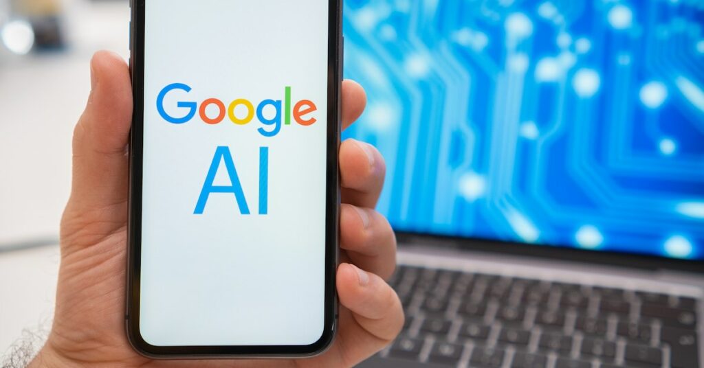 The smartphone screen held by a man shows a Google AI neural network logo, while in the background, a laptop displays a splash screen with a neuronet.